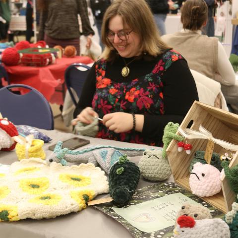 Student knitting at table with other knitted goods.