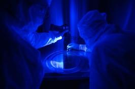 Two people in white Tyvek suits examine a thin flat disk under a blue ultraviolet light that detects dust.
