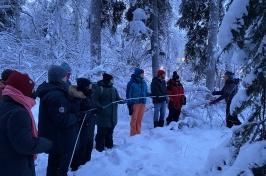 People bundled up in winter gear hold a long white tube to measure soil frost amidst a snowy forest. 